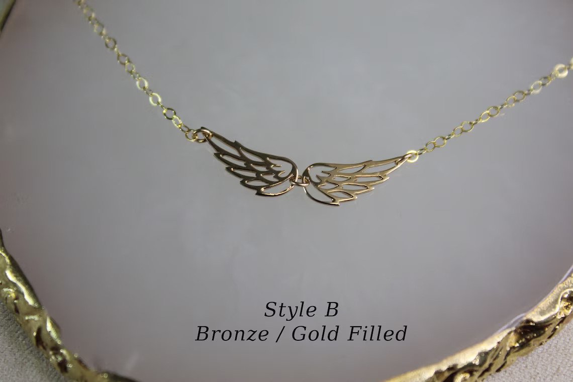 Gold filled necklace with two small angel wing charms is displayed on a white marble background.