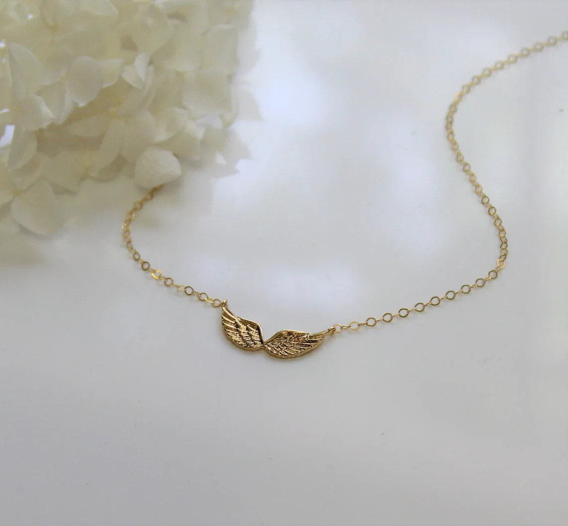 Angel wing charm attached to necklace chain in a gold finish is laying on a shiny surface with decorative white flowers in the background.
