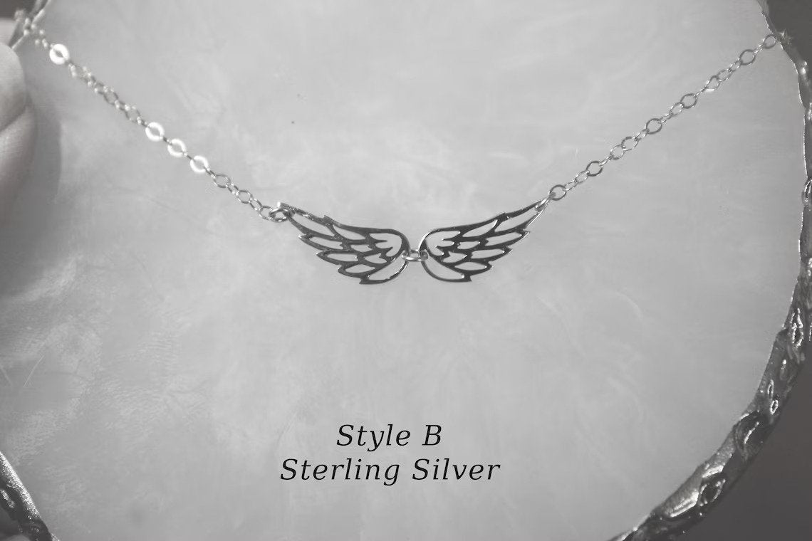 Two angel wings are connected on a necklace in a sterling silver finish and displayed against a plain marble surface.
