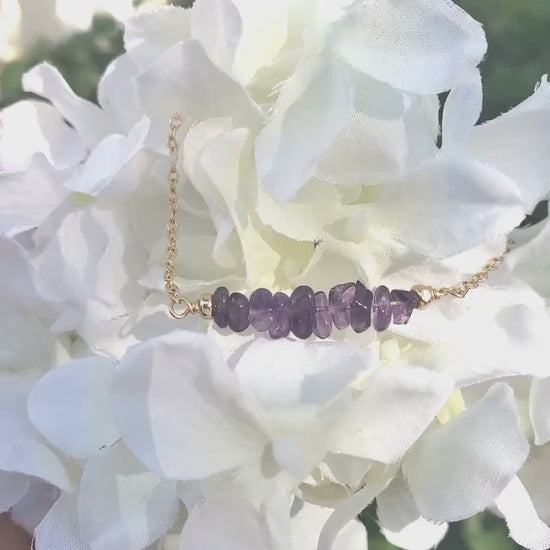 Natural amethyst gemstones wire wrapped onto a gold filled necklace hang on a synthetic white flower and glisten in the sun.