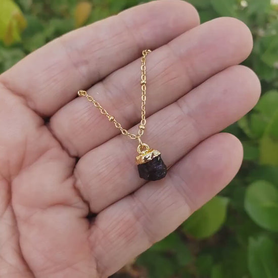 Raw amethyst gemstone pendant is electroplated with genuine gold and hangs on a gold filled satellite chain necklace which is being held in a womans hand and glistens inthe sun.