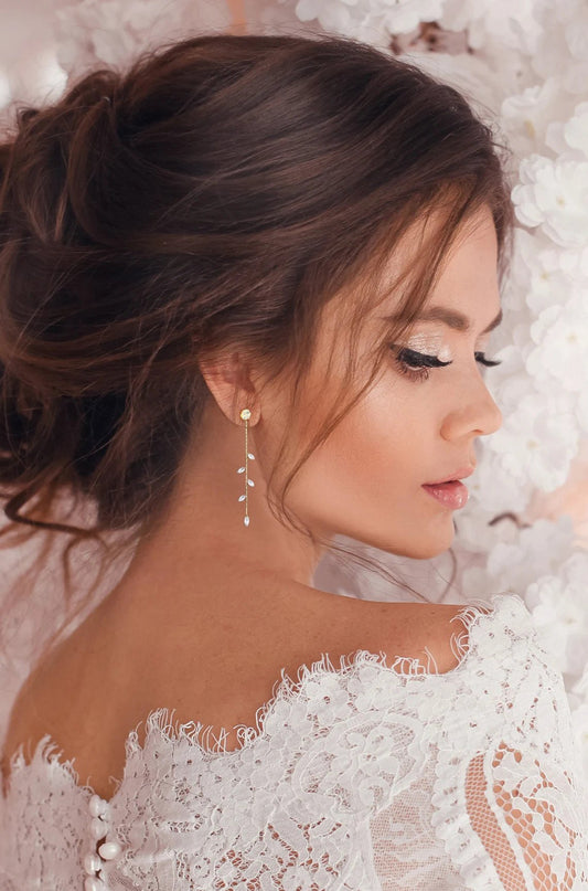 Bridal model in a lacey of the shoulder dress and a fancy up hair style  stands in front of a wall of white flowers and displays her crystal dangle earrings.