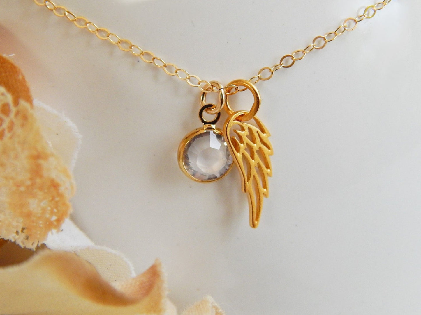 Angel wing charm hanging next to a swarovski birthstone charm on a gold necklace chain.