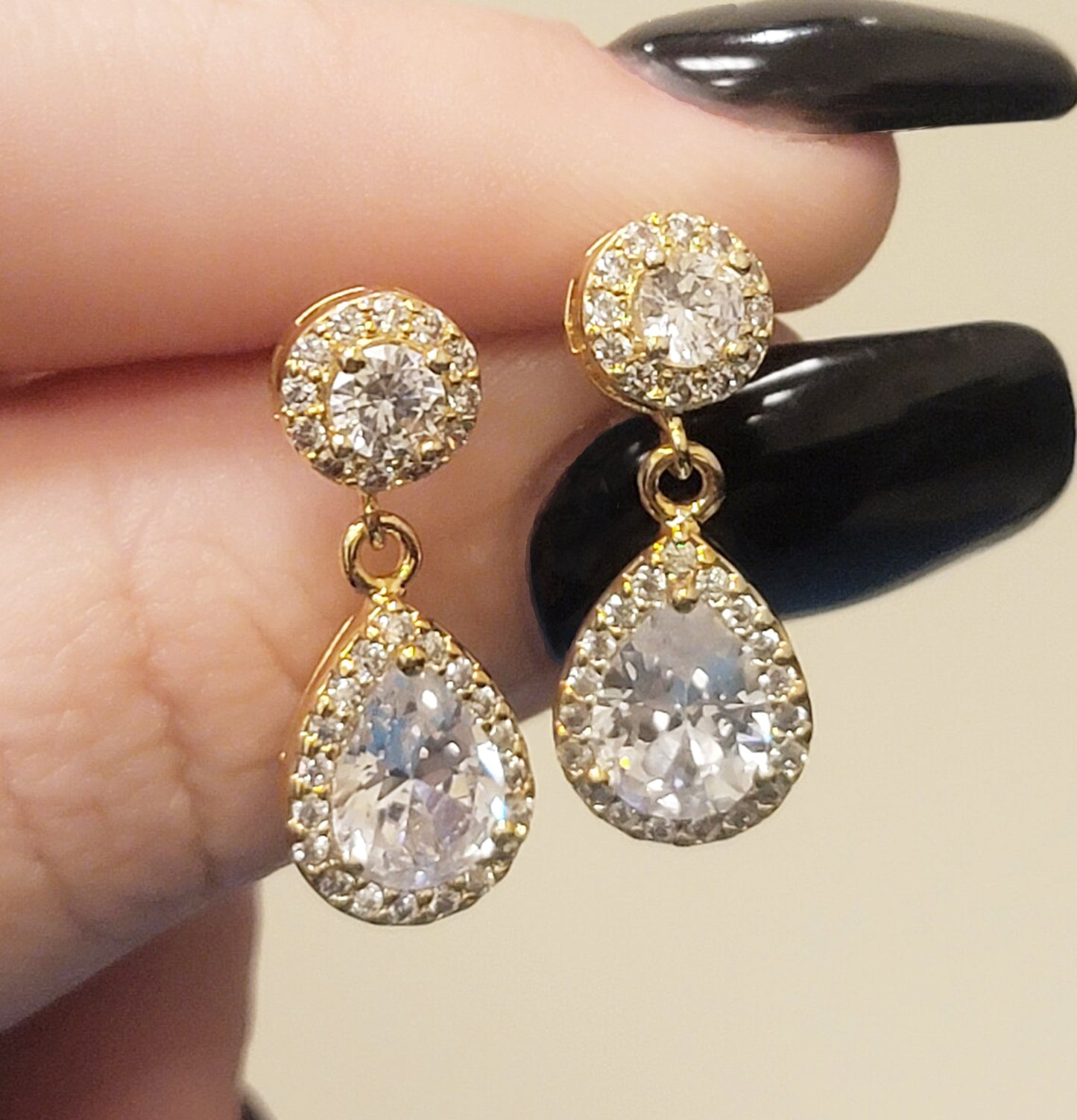 Gold filled special event prom and wedding earrings with many cubic zirconias and large teardrop diamond and held between  woman's thumb and forefinger with black manicure.