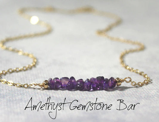 Natural amethyst gemstone chips beaded onto a gold filled wire to create a gemstone bar necklace laying on a textured gray surface.