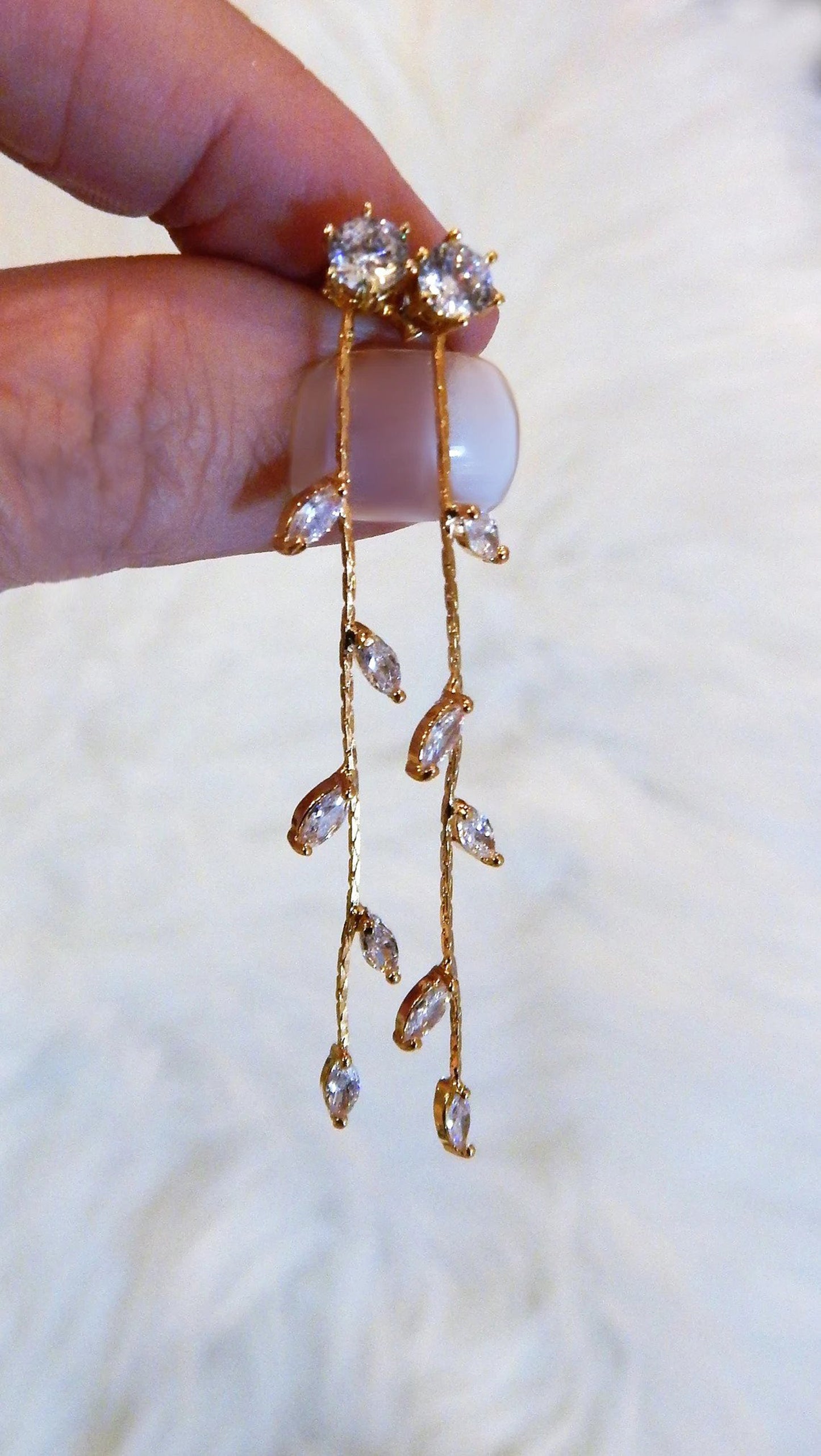 Two earrings with a 3 inch drop and diamond cubic zirconias are held between a womans fingers against a white background.