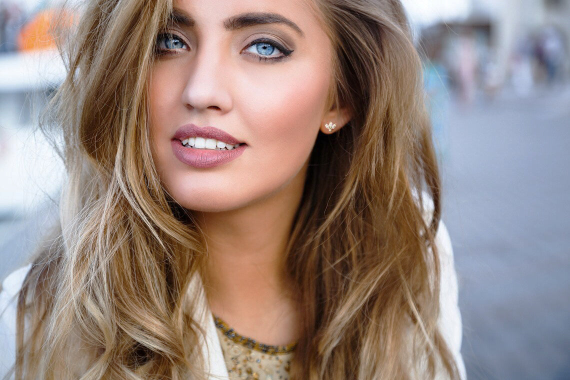 Portrait of a fashion model with blue eyes and dirty blonde wavy hair is wearing a simple gold diamond earring stud with a blurry city background behind her.