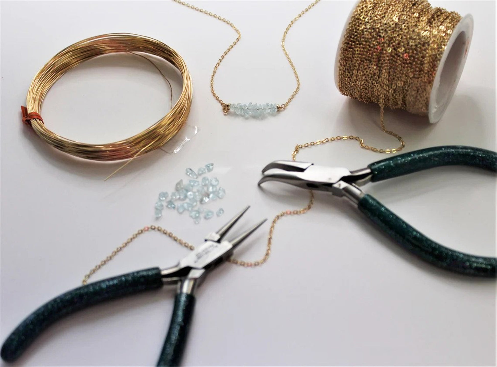 Custom jewelers tools and supplies including gold filled necklace chain, wire wrapping, natural gemstone pieces, jewelers pliers, and a partially complete gemstone necklace lay on a flat white surface.