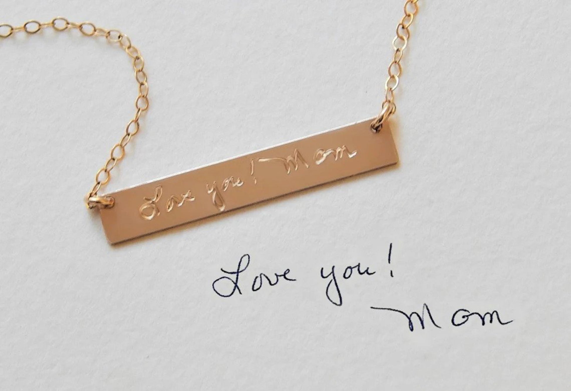Custom gold filled keepsake jewelry by Gilded Sapphire shows engraved gold bar necklace with personalized message etched on its surface laying on a piece of paper with the same handwritten note engraved on the bar.