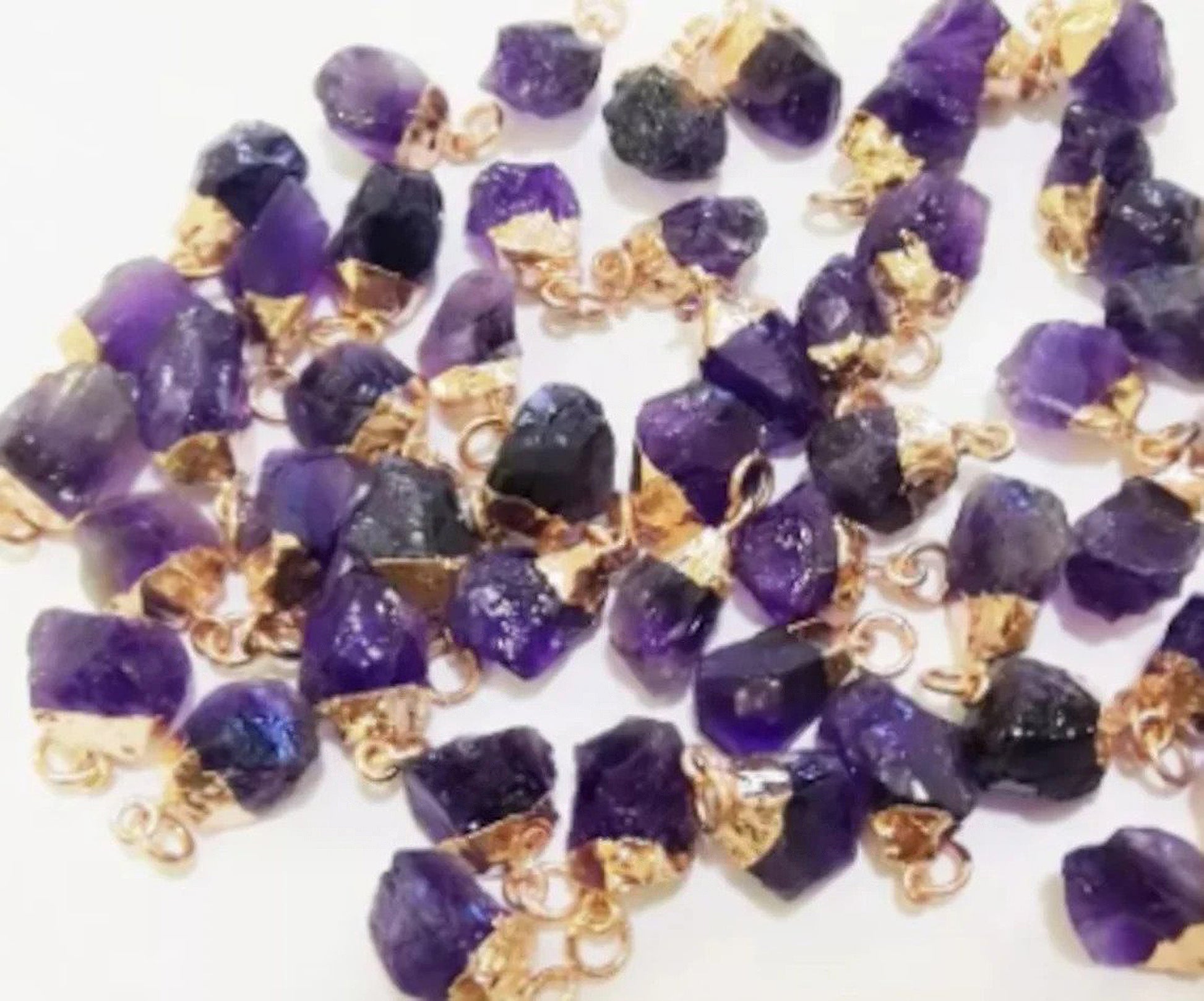Small amethyst crystals in their raw and unpolished form have been electroplated with gold to create dainty jewelry pendants which lay in a pile together on top of a plain white surface.