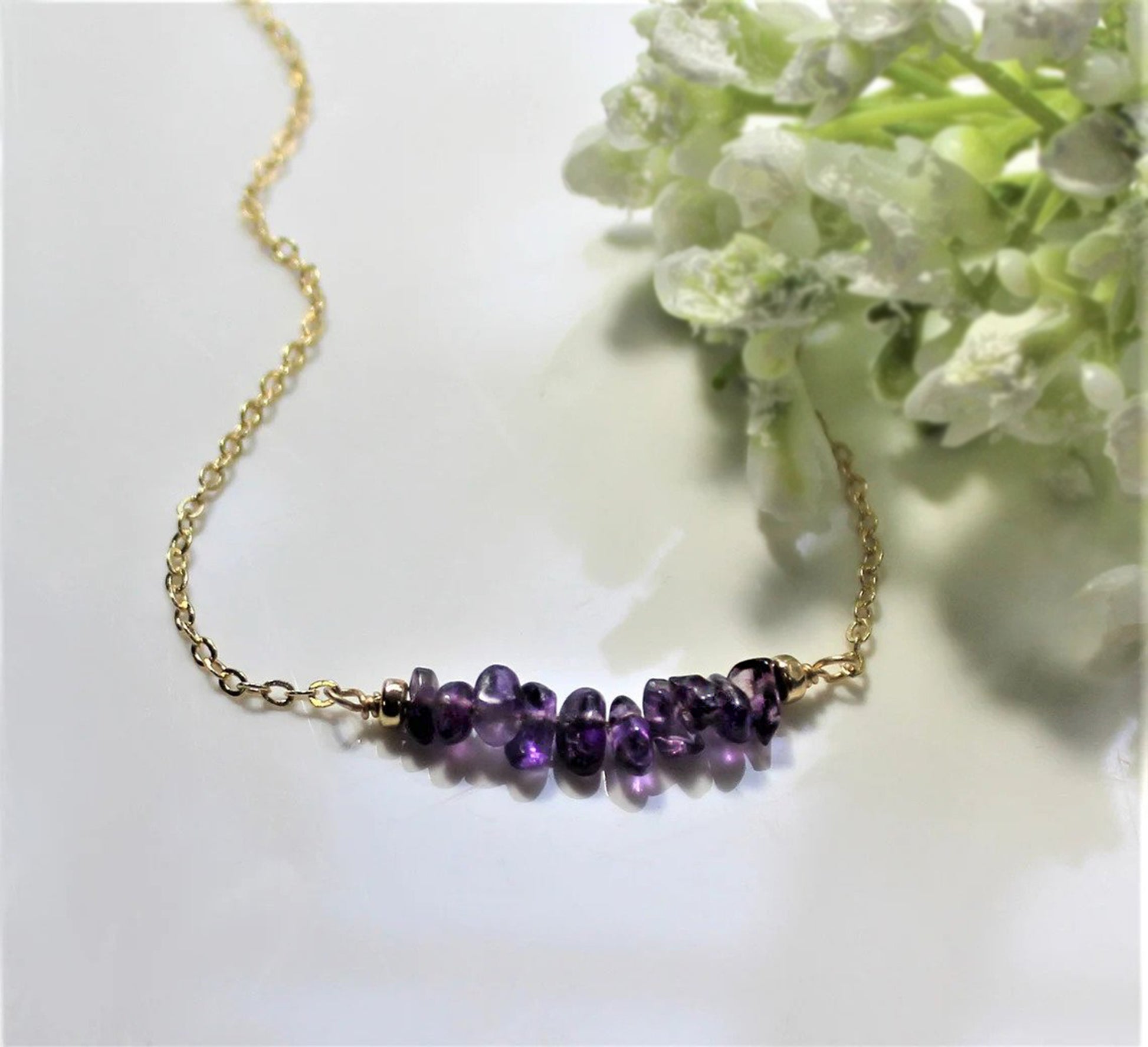 Natural amethyst gemstone chips beaded onto jewelers wire creates a handmade birthstone bar necklace complete with gold filled chain laying on a plain white display surface and decorative white and green flowers in the background.