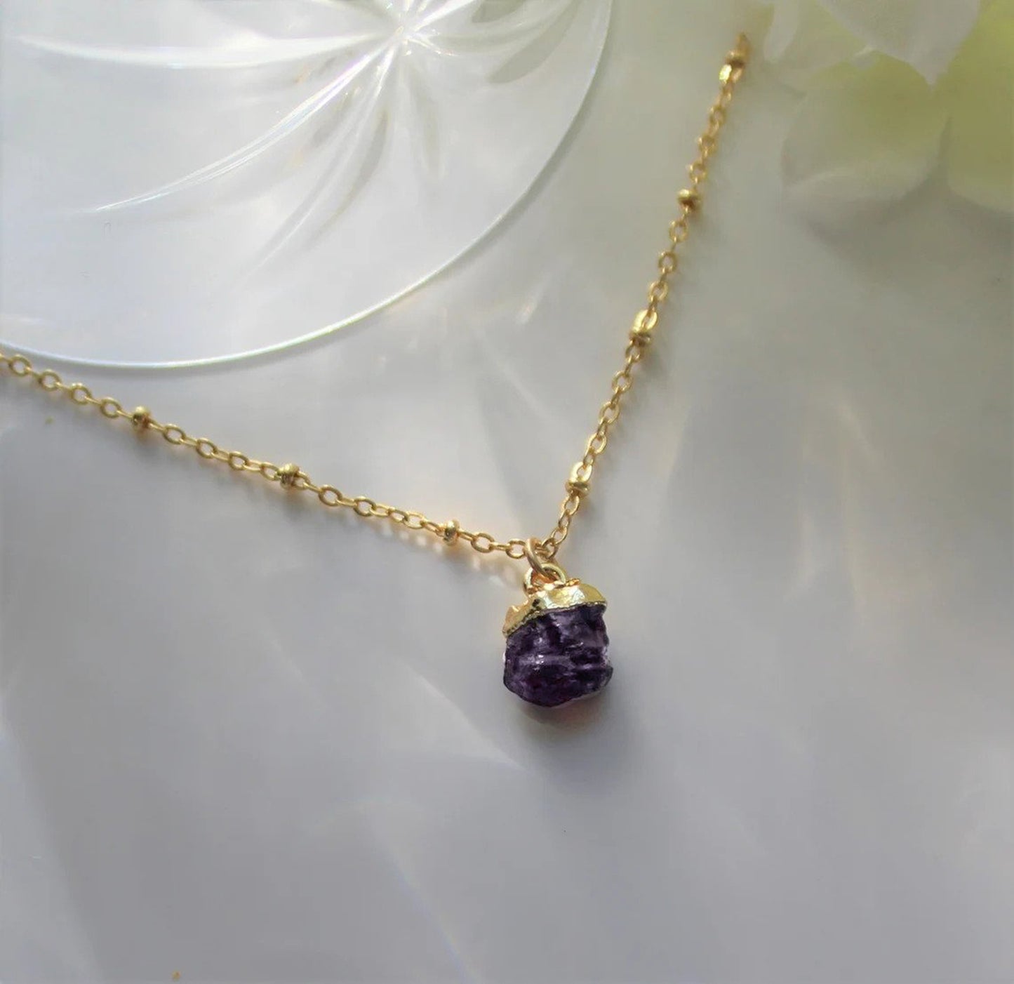 Natural purple amethyst gemstone has been made into a gold pendant and is featured on a gold filled satellite necklace chain and is displayed on a pale green surface with many light reflections.