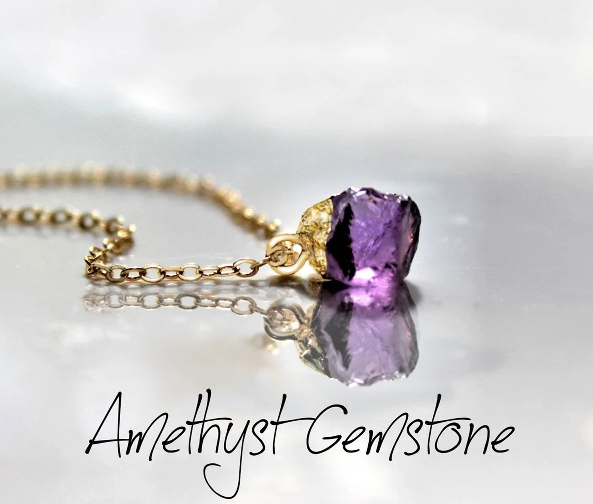 Raw amethyst crystal with a gold electroplated top creates a dainty pendant which is placed on a gold filled necklace chain and laying on a reflective surface to display both the gemstone and its mirror image.