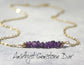 Natural amethyst gemstone chips beaded onto a gold filled wire to create a gemstone bar necklace laying on a textured gray surface.