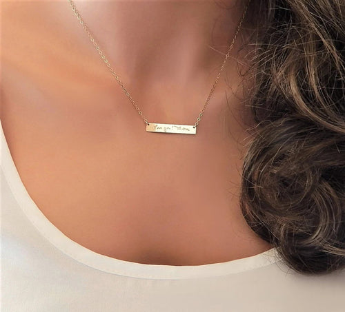A mannequin torso wearing a white shirt and gold necklace. The necklace has a gold horizontal bar that has been engraved with custom text in personal handwriting.