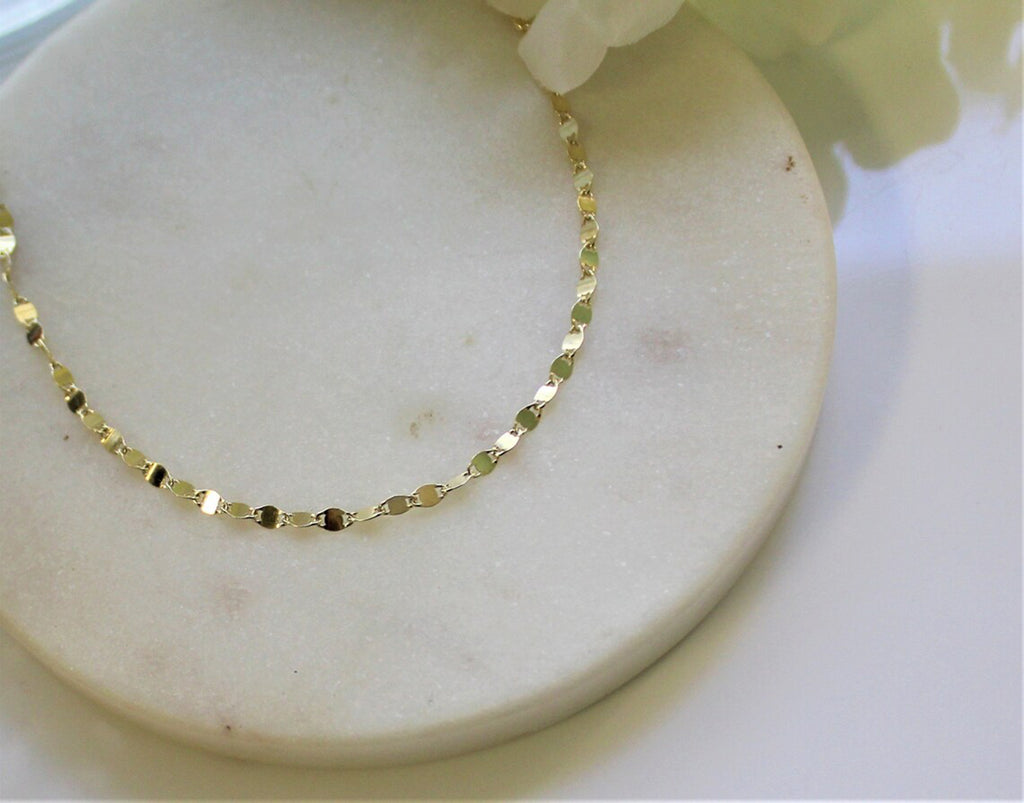 Solid gold sequin necklace chain laying on a white marble coaster with white flowers showcasing Gilded Sapphire's custom jewelry.